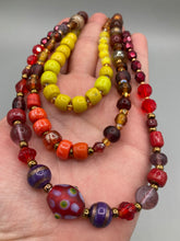 Load image into Gallery viewer, This necklace consists of three strands with a multitude of shades ranging from red to orange, gold, and amber, providing a captivating visual combination. Crafted with a combination of glass, ceramic and metal beads, this eye-catching piece of jewelry radiates power and strength. Measurements: Necklace is 16” in length, can be adjusted up to 17 1/2"