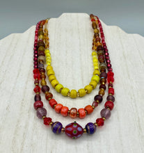 Load image into Gallery viewer, This necklace consists of three strands with a multitude of shades ranging from red to orange, gold, and amber, providing a captivating visual combination. Crafted with a combination of glass, ceramic and metal beads, this eye-catching piece of jewelry radiates power and strength. Measurements: Necklace is 16” in length, can be adjusted up to 17 1/2"
