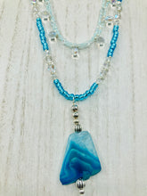 Load image into Gallery viewer, This exquisite necklace features a vibrant Sea Blue Agate Natural Stone Pendant and two strands of multi-colored glass beaded necklace. An adjustable collar-style strand contains small, light blue beads and lots of iridescent double-hole beads for shimmer and sparkle. The longer strand features larger blue glass beads interspersed with clear and silver beads for a sophisticated, draped look. It's finished with a set of dainty matching earrings for a complete look. Necklace is 14”adjustable up to 18"