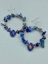 Load image into Gallery viewer, Featuring natural lapis stone chips and royal blue glass beads with golden accents, these hoop earrings complete any look with a dark blue pressed glass leaf charm boasting an iridescent shimmer with hints of gold, purple, blue and pink. Measurements: 1 3/4" length
