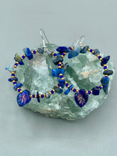 Load image into Gallery viewer, Featuring natural lapis stone chips and royal blue glass beads with golden accents, these hoop earrings complete any look with a dark blue pressed glass leaf charm boasting an iridescent shimmer with hints of gold, purple, blue and pink. Measurements: 1 3/4" length