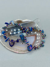 Load image into Gallery viewer, Featuring natural lapis stone chips and royal blue glass beads with golden accents, these hoop earrings complete any look with a dark blue pressed glass leaf charm boasting an iridescent shimmer with hints of gold, purple, blue and pink. Measurements: 1 3/4" length