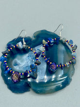 Load image into Gallery viewer, Featuring natural lapis stone chips and royal blue glass beads with golden accents, these hoop earrings complete any look with a dark blue pressed glass leaf charm boasting an iridescent shimmer with hints of gold, purple, blue and pink. Measurements: 1 3/4" length