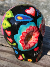 Load image into Gallery viewer, Inspired by nature, this ceramic skull statuette is full of vibrant color and intricate details. The primary color is black with multi-color floral motifs that are painted across the skull, along with hearts and leaves and lively colorful accents around the entire skull.
