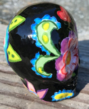 Load image into Gallery viewer, Inspired by nature, this ceramic skull statuette is full of vibrant color and intricate details. The primary color is black with multi-color floral motifs that are painted across the skull, along with hearts and leaves and lively colorful accents around the entire skull.