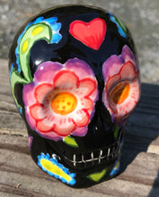 Load image into Gallery viewer, Inspired by nature, this ceramic skull statuette is full of vibrant color and intricate details. The primary color is black with multi-color floral motifs that are painted across the skull, along with hearts and leaves and lively colorful accents around the entire skull.