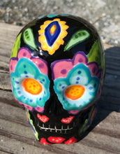 Load image into Gallery viewer, This black ceramic sugar skull, inspired by nature, is adorned with hand-painted floral motifs, hearts, leaves and colorful accents. Its vibrant style and intricate details create a stunning Dia De Los Muertos calavera. Perfect for creating ofrendas to celebrate the Day of the Dead. Measurements: H 2 3/4” x W 2” x D 2”