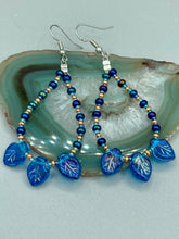 Load image into Gallery viewer, These striking Royal Blue Pressed Glass Leaf Beaded Hoop Earrings feature a shimmery finish with a hint of gold, purple, blue and pink. Gold, dark and light blue glass beads complete the piece. Measurements: 2 1/2" length