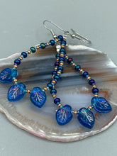 Load image into Gallery viewer, These striking Royal Blue Pressed Glass Leaf Beaded Hoop Earrings feature a shimmery finish with a hint of gold, purple, blue and pink. Gold, dark and light blue glass beads complete the piece. Measurements: 2 1/2" length