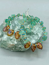 Load image into Gallery viewer, These Aventurine Stone Chip Beaded Hoop Earrings feature natural aventurine stone chips, turquoise, and gold glass beads. The amber-tone pressed glass leaf charm has an iridescent shimmer finish, with subtle accents of gold, purple, blue, and pink. Measurements: 2" length