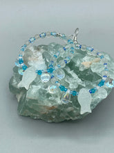 Load image into Gallery viewer, This stylish set of earrings feature milky white fish-shaped glass beads, complemented by light blue and iridescent teardrop glass beads. The fish-shaped glass beads feature intricate details, including fins and an eye. Measurements: 1 7/8” in length