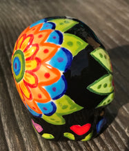 Load image into Gallery viewer, Inspired by nature, this ceramic skull statuette is full of vibrant color and intricate details. The primary color is black with multi-color floral motifs that are painted across the skull, along with hearts and leaves and lively colorful accents around the entire skull.