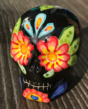 Load image into Gallery viewer, Inspired by nature, this ceramic skull statuette is full of vibrant color and intricate details. The primary color is black with multi-color floral motifs that are painted across the skull, along with hearts and leaves and lively colorful accents around the entire skull.