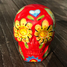 Load image into Gallery viewer, This ceramic skull statuette is a stunning piece of art inspired by nature. Its vibrant red hue is decorated with multi-color floral motifs, hearts, leaves and colorful accents, creating a beautiful piece of hand-painted, Day of the Dead-themed art. Measurements: H 2 3/4” x W 2” x D 2”