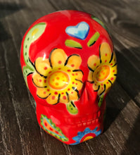 Load image into Gallery viewer, This ceramic skull statuette is a stunning piece of art inspired by nature. Its vibrant red hue is decorated with multi-color floral motifs, hearts, leaves and colorful accents, creating a beautiful piece of hand-painted, Day of the Dead-themed art. Measurements: H 2 3/4” x W 2” x D 2”
