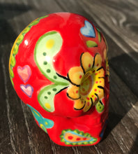 Load image into Gallery viewer, This ceramic skull statuette is a stunning piece of art inspired by nature. Its vibrant red hue is decorated with multi-color floral motifs, hearts, leaves and colorful accents, creating a beautiful piece of hand-painted, Day of the Dead-themed art. Measurements: H 2 3/4” x W 2” x D 2”