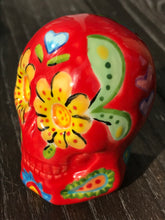 Load image into Gallery viewer, This ceramic skull statuette is a stunning piece of art inspired by nature. Its vibrant red hue is decorated with multi-color floral motifs, hearts, leaves and colorful accents, creating a beautiful piece of hand-painted, Day of the Dead-themed art. Measurements: H 2 3/4” x W 2” x D 2”