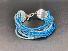 Load image into Gallery viewer, This bracelet is a chic, minimalistic design with nine beaded strands that feature various blue and silver glass beads. It measures 7 1/2" in length and has been crafted with inspiration from the ocean blue waves.