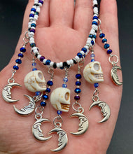 Load image into Gallery viewer, Howlite Skull Beads And Smiling Crescent Moon Charms Choker Style Glass Beaded Necklace. Two beaded stands that are primarily white and added purple glass beads that have hues of blue and gold. I wanted to incorporate dark and white glass bead colors to create a simple contrast. Large amount of dangling skulls and their smiling crescent moons add a lot of movement. Necklace is 16”adjustable up to 18" in length - hanging howlite skull with crescent moons are 2” Height