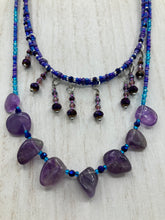 Load image into Gallery viewer, Three beaded strands using a wide range of size, shape, and color glass beads. The first two shorter stands are primarily purple glass beads that have tiny shimmery hues of blue, pink and gold. I added a series of dangling glass beads along this necklace to give this collar some movement and make it stand out even more. The third and longer strand contains a large amount of amethyst nugget beads. Large necklace hangs beautifully across your neck and chest. Necklace is 16” in adjustable up to 18" in