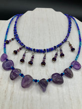 Load image into Gallery viewer, Three beaded strands using a wide range of size, shape, and color glass beads. The first two shorter stands are primarily purple glass beads that have tiny shimmery hues of blue, pink and gold. I added a series of dangling glass beads along this necklace to give this collar some movement and make it stand out even more. The third and longer strand contains a large amount of amethyst nugget beads. Large necklace hangs beautifully across your neck and chest. Necklace is 16” in adjustable up to 18" in