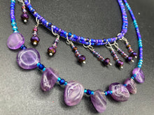 Load image into Gallery viewer, Three beaded strands using a wide range of size, shape, and color glass beads. The first two shorter stands are primarily purple glass beads that have tiny shimmery hues of blue, pink and gold. I added a series of dangling glass beads along this necklace to give this collar some movement and make it stand out even more. The third and longer strand contains a large amount of amethyst nugget beads. Large necklace hangs beautifully across your neck and chest. Necklace is 16” in adjustable up to 18" in