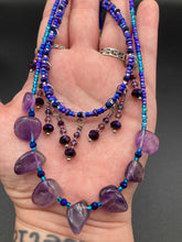 Load image into Gallery viewer, Three beaded strands using a wide range of size, shape, and color glass beads. The first two shorter stands are primarily purple glass beads that have tiny shimmery hues of blue, pink and gold. I added a series of dangling glass beads along this necklace to give this collar some movement and make it stand out even more. The third and longer strand contains a large amount of amethyst nugget beads. Large necklace hangs beautifully across your neck and chest. Necklace is 16” in adjustable up to 18" in