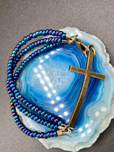 Load image into Gallery viewer, For Positivity and Faith, Shimmery Blue Glass Beaded Bracelet With Curved Horizontal Brass Cross. This bracelet has a large brass cross that is slightly curved to be comfortably worm around your wrist. I created three glass beaded strands that have a wide variety of blue, purple, green, and gold tones. Measurements: 7 1/2" in length, brass cross measures 2" in length