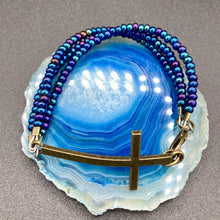 Load image into Gallery viewer, For Positivity and Faith, Shimmery Blue Glass Beaded Bracelet With Curved Horizontal Brass Cross. This bracelet has a large brass cross that is slightly curved to be comfortably worm around your wrist. I created three glass beaded strands that have a wide variety of blue, purple, green, and gold tones. Measurements: 7 1/2" in length, brass cross measures 2" in length