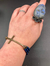 Load image into Gallery viewer, For Positivity and Faith, Shimmery Blue Glass Beaded Bracelet With Curved Horizontal Brass Cross. This bracelet has a large brass cross that is slightly curved to be comfortably worm around your wrist. I created three glass beaded strands that have a wide variety of blue, purple, green, and gold tones. Measurements: 7 1/2" in length, brass cross measures 2" in length