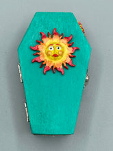 Load image into Gallery viewer, This coffin-shaped box features a bright turquoise exterior, with a bold yellow, orange and red sun adorning the center. Complemented with a shimmery rose-gold ribbon and flower, the inside is painted in a vibrant orange hue with turquoise crescent moon detailing and traditional Mexican-themed fabric. Coffin Dimensions: Length: 4 1/2 Inches; Width: 2 1/2 inches; Height: 1 5/8 Inches
