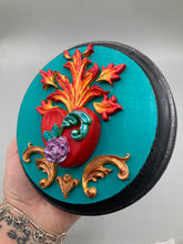 Load image into Gallery viewer, This Sacred Heart Wall Art Decor is the perfect way to enhance your home with an artistic flair. This beautiful artwork features a round bright turquoise wooden frame with a hand-crafted bright red heart. The heart is detailed with light purple and vibrant green leaves, with a vibrant fire embellishment over the center. Gold decorative embellishments at the sides of the heart add a luxurious touch. Diameter 4"