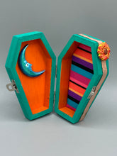 Load image into Gallery viewer, This coffin-shaped box features a bright turquoise exterior, with a bold yellow, orange and red sun adorning the center. Complemented with a shimmery rose-gold ribbon and flower, the inside is painted in a vibrant orange hue with turquoise crescent moon detailing and traditional Mexican-themed fabric. Coffin Dimensions: Length: 4 1/2 Inches; Width: 2 1/2 inches; Height: 1 5/8 Inches