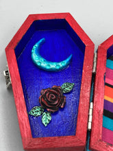 Load image into Gallery viewer, This coffin-shaped box features a bright red exterior, with a bold yellow, orange and red sun adorning the center. Complemented with a shimmery turquoise-blue ribbon and flower, the inside is painted in a dark royal blue hue with turquoise crescent moon detailing and traditional Mexican-themed fabric. Coffin Dimensions: Length: 4 1/2 Inches; Width: 2 1/2 inches; Height: 1 5/8 Inches
