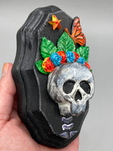 Load image into Gallery viewer, Showcasing a vibrant Frida Kahlo inspired calavera with a dark wood frame, this Day of the Dead wall decor piece is perfect for adding a traditional Mexican folk art touch to any home. Featuring an intricately detailed clay calavera in the style of Frida Kahlo, this Day of the Dead wall art decor is sure to be the centerpiece of any home décor. The calavera is wearing Frida's iconic crown of flowers and a monarch butterfly, ensuring it will bring both a unique and inspiring look to any wall.
