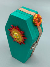 Load image into Gallery viewer, This coffin-shaped box features a bright turquoise exterior, with a bold yellow, orange and red sun adorning the center. Complemented with a shimmery rose-gold ribbon and flower, the inside is painted in a vibrant orange hue with turquoise crescent moon detailing and traditional Mexican-themed fabric. Coffin Dimensions: Length: 4 1/2 Inches; Width: 2 1/2 inches; Height: 1 5/8 Inches