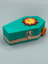 Load image into Gallery viewer, This coffin-shaped box features a bright turquoise exterior, with a bold yellow, orange and red sun adorning the center. Complemented with a shimmery rose-gold ribbon and flower, the inside is painted in a vibrant orange hue with turquoise crescent moon detailing and traditional Mexican-themed fabric. Coffin Dimensions: Length: 4 1/2 Inches; Width: 2 1/2 inches; Height: 1 5/8 Inches