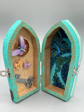 Load image into Gallery viewer, This Whimsical Fairy Wood Coffin Box is a truly unique and magical piece of home decor. With its vibrant turquoise exterior and eye-catching mini door knob, this Steampunk-inspired box is sure to be a conversation starter. Inside, the glittery gold interior and colorful butterfly detailing will bring a touch of fantasy to any room. The silver key and accompanying iridescent crystals complete the mystical and pastel goth look!