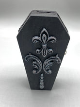 Load image into Gallery viewer, This vintage-inspired coffin ring box will make a spooky yet cute addition to any jewelry collection. The sleek black box has eye-catching black and gray embellishments, while the interior is beautifully decorated with a vivid turquoise blue fabric and a highly detailed black and silver cross with dark red roses. Function and style meet with this unique and stylish piece.