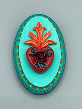 Load image into Gallery viewer, This Sacred Heart Wall Art Decor is the perfect way to enhance your home with an artistic flair. This beautiful artwork features a bright turquoise wooden frame with a hand-crafted bright red heart. The heart is detailed with dark red rose and vibrant green leaves, with a vibrant fire embellishment over the center. Gold decorative embellishments at the sides of the heart add a luxurious touch. H 5 3/8" x W 3 3/8” x D 1 1/2"