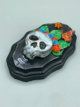 Load image into Gallery viewer, Showcasing a vibrant Frida Kahlo inspired calavera with a dark wood frame, this Day of the Dead wall decor piece is perfect for adding a traditional Mexican folk art touch to any home. Featuring an intricately detailed clay calavera in the style of Frida Kahlo, this Day of the Dead wall art decor is sure to be the centerpiece of any home décor. The calavera is wearing Frida's iconic crown of flowers and a monarch butterfly, ensuring it will bring both a unique and inspiring look to any wall.