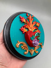 Load image into Gallery viewer, This Sacred Heart Wall Art Decor is the perfect way to enhance your home with an artistic flair. This beautiful artwork features a round bright turquoise wooden frame with a hand-crafted bright red heart. The heart is detailed with light purple and vibrant green leaves, with a vibrant fire embellishment over the center. Gold decorative embellishments at the sides of the heart add a luxurious touch. Diameter 4"