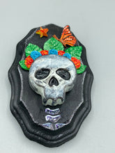 Load image into Gallery viewer, Showcasing a vibrant Frida Kahlo inspired calavera with a dark wood frame, this Day of the Dead wall decor piece is perfect for adding a traditional Mexican folk art touch to any home. Featuring an intricately detailed clay calavera in the style of Frida Kahlo, this Day of the Dead wall art decor is sure to be the centerpiece of any home décor. The calavera is wearing Frida's iconic crown of flowers and a monarch butterfly, ensuring it will bring both a unique and inspiring look to any wall.