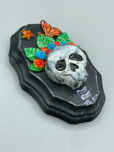 Load image into Gallery viewer, Showcasing a vibrant Frida Kahlo inspired calavera with a dark wood frame, this Day of the Dead wall decor piece is perfect for adding a traditional Mexican folk art touch to any home. Featuring an intricately detailed clay calavera in the style of Frida Kahlo, this Day of the Dead wall art decor is sure to be the centerpiece of any home décor. The calavera is wearing Frida's iconic crown of flowers and a monarch butterfly, ensuring it will bring both a unique and inspiring look to any wall.