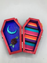 Load image into Gallery viewer, This coffin-shaped box features a bright red exterior, with a bold yellow, orange and red sun adorning the center. Complemented with a shimmery turquoise-blue ribbon and flower, the inside is painted in a dark royal blue hue with turquoise crescent moon detailing and traditional Mexican-themed fabric. Coffin Dimensions: Length: 4 1/2 Inches; Width: 2 1/2 inches; Height: 1 5/8 Inches