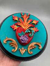 Load image into Gallery viewer, This Sacred Heart Wall Art Decor is the perfect way to enhance your home with an artistic flair. This beautiful artwork features a round bright turquoise wooden frame with a hand-crafted bright red heart. The heart is detailed with light purple and vibrant green leaves, with a vibrant fire embellishment over the center. Gold decorative embellishments at the sides of the heart add a luxurious touch. Diameter 4"