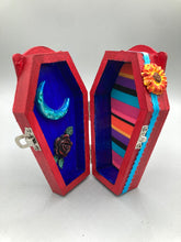Load image into Gallery viewer, This coffin-shaped box features a bright red exterior, with a bold yellow, orange and red sun adorning the center. Complemented with a shimmery turquoise-blue ribbon and flower, the inside is painted in a dark royal blue hue with turquoise crescent moon detailing and traditional Mexican-themed fabric. Coffin Dimensions: Length: 4 1/2 Inches; Width: 2 1/2 inches; Height: 1 5/8 Inches