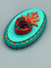 Load image into Gallery viewer, This Sacred Heart Wall Art Decor is the perfect way to enhance your home with an artistic flair. This beautiful artwork features a bright turquoise wooden frame with a hand-crafted bright red heart. The heart is detailed with dark red rose and vibrant green leaves, with a vibrant fire embellishment over the center. Gold decorative embellishments at the sides of the heart add a luxurious touch. H 5 3/8" x W 3 3/8” x D 1 1/2"