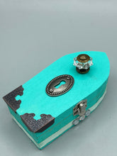 Load image into Gallery viewer, This Whimsical Fairy Wood Coffin Box is a truly unique and magical piece of home decor. With its vibrant turquoise exterior and eye-catching mini door knob, this Steampunk-inspired box is sure to be a conversation starter. Inside, the glittery gold interior and colorful butterfly detailing will bring a touch of fantasy to any room. The silver key and accompanying iridescent crystals complete the mystical and pastel goth look!