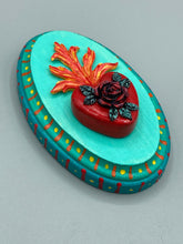 Load image into Gallery viewer, This Sacred Heart Wall Art Decor is the perfect way to enhance your home with an artistic flair. This beautiful artwork features a bright turquoise wooden frame with a hand-crafted bright red heart. The heart is detailed with dark red rose and vibrant green leaves, with a vibrant fire embellishment over the center. Gold decorative embellishments at the sides of the heart add a luxurious touch. H 5 3/8" x W 3 3/8” x D 1 1/2"